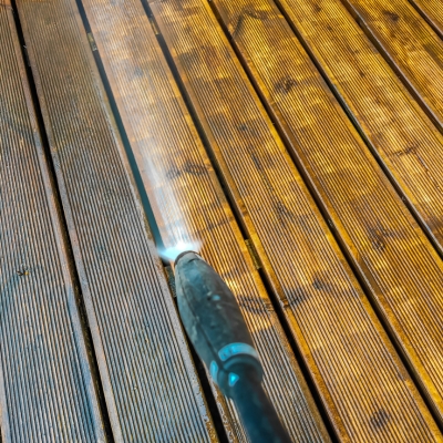 A close-up of a pressure washer cleaning a wooden deck with visible dirt and grime being removed.