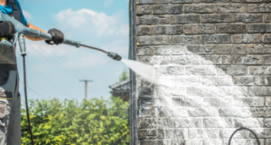 A worker using a pressure washer to clean a dark grey brick wall on a sunny day.