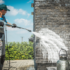 A worker using a pressure washer to clean a dark grey brick wall on a sunny day.