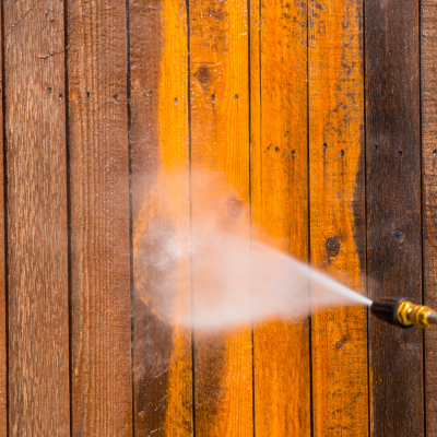 A pressure washer cleaning a weathered wooden fence, revealing a vibrant wood surface beneath.