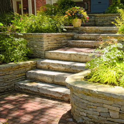 Stone steps surrounded by garden plants, leading to a house entrance, with brick paving and stone walls.