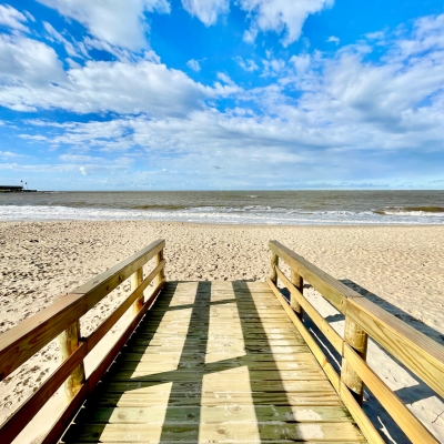 A wooden decking walkway leading to a sandy beach and calm sea under a blue sky.