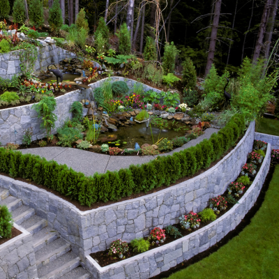 A multi-level terraced landscape with stone walls, greenery, flower beds, and a pond in a forest setting.