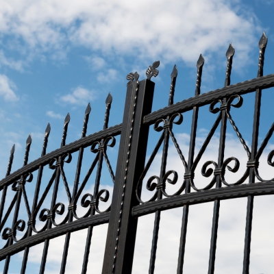 A black wrought iron gate with intricate detailing, set against a bright blue sky with soft clouds.