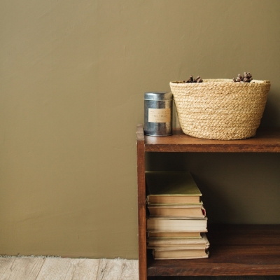 A warm olive green-painted wall with a wooden shelf, stacked books, a woven basket, and a tin container.