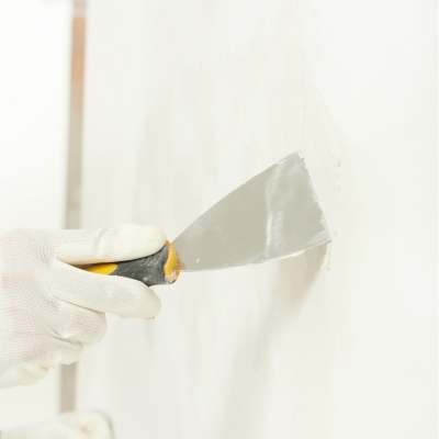 A person wearing gloves uses a putty knife to apply filler on a smooth wall.