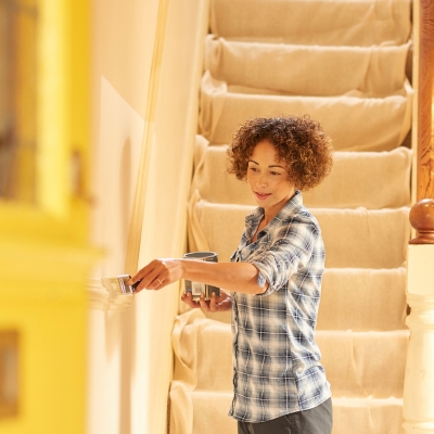 A woman in a plaid shirt painting a hallway wall in a soft, neutral shade with a paintbrush.