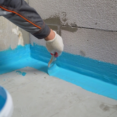 A hand applying blue waterproofing membrane to the floor and wall junction of a wet room.