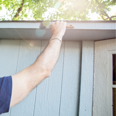 A person painting the exterior of a wooden shed in a muted grey tone with a paintbrush.