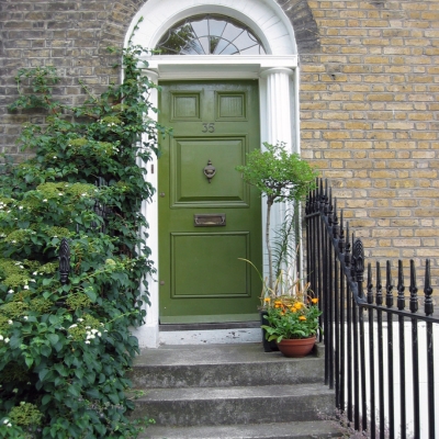 A deep olive green front door on a traditional brick townhouse, surrounded by plants and black railings.