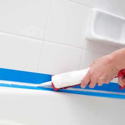 A person applying white waterproof caulk along the edge of a bathtub using a caulking gun.