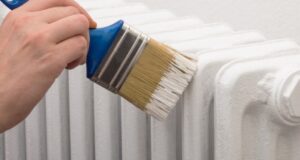 A person using a paintbrush to apply white paint to a metal radiator.