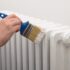 A person using a paintbrush to apply white paint to a metal radiator.