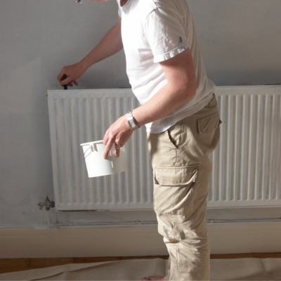 A person standing in front of a radiator, holding a paint tin and brush, ready to paint.