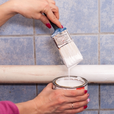 A person dipping a brush into white radiator paint, preparing to coat heating pipes.