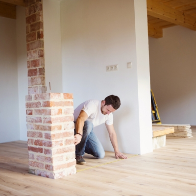 A man crouching on a wooden floor using a tape measure to measure around a brick column.