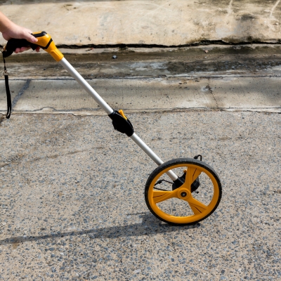 A person using a yellow measuring wheel on an outdoor concrete surface.